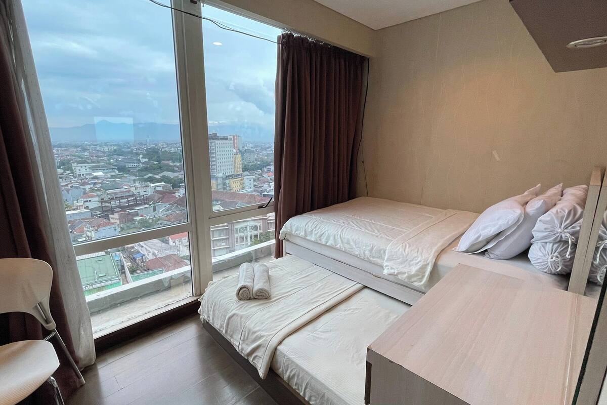 A hotel room with two beds, a large window with a city view, a chair, and a wooden desk.