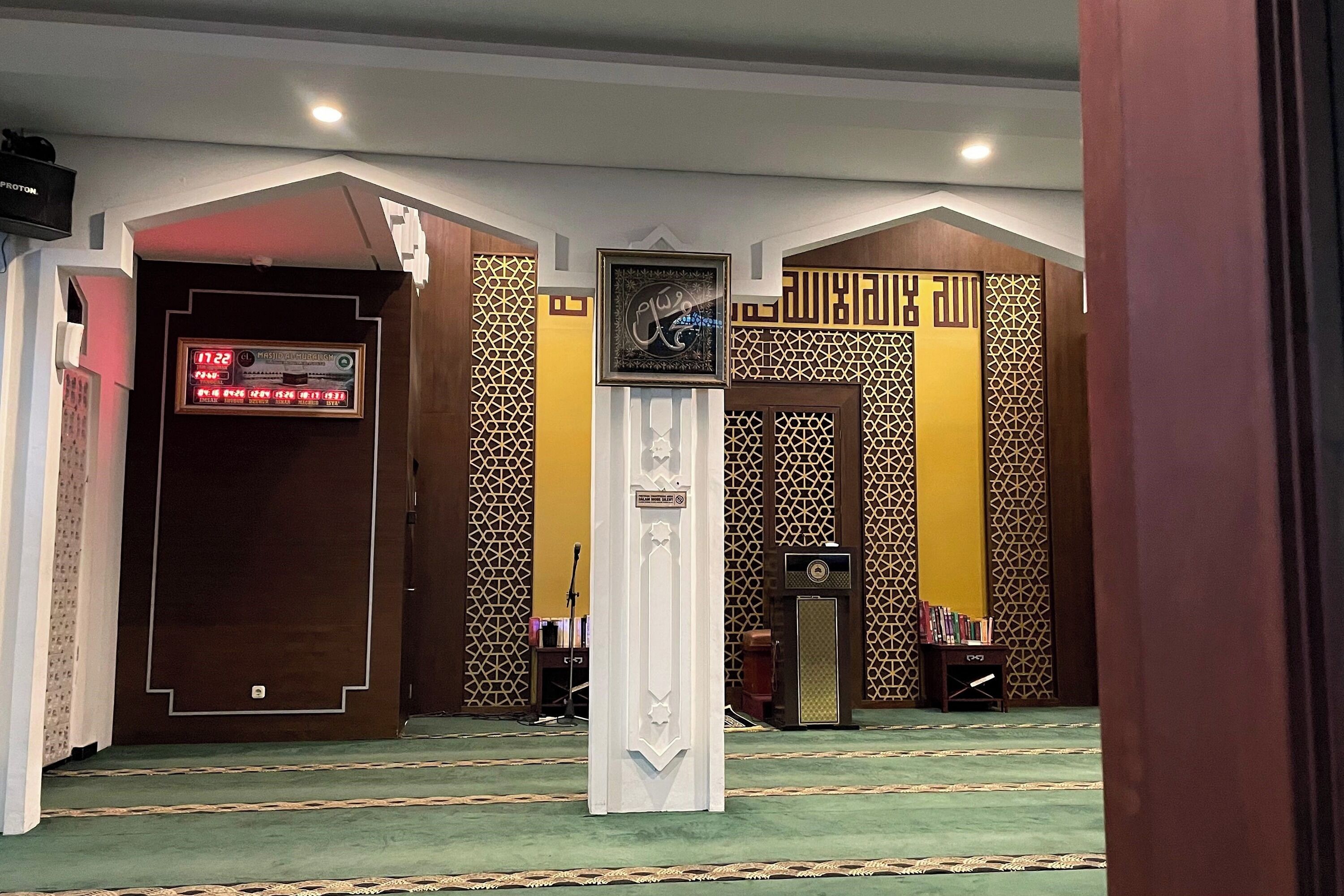 A prayer hall with a green carpet, wooden walls with geometric patterns, a white pillar, and a digital clock.