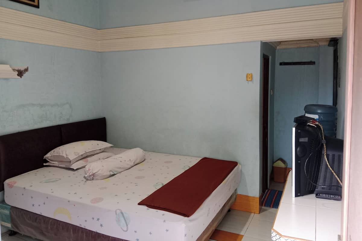 Een slaapkamer met een bed, kussens en een deken. Er staat een waterkoeler en een klein tafeltje met een stoel.