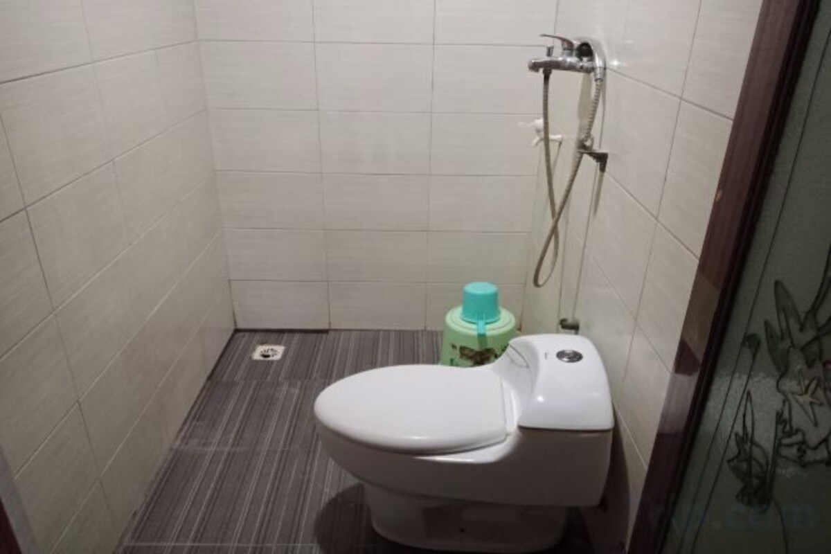 Une petite salle de bain carrelée, avec des toilettes, une douche et une bouteille verte posée sur le sol.
