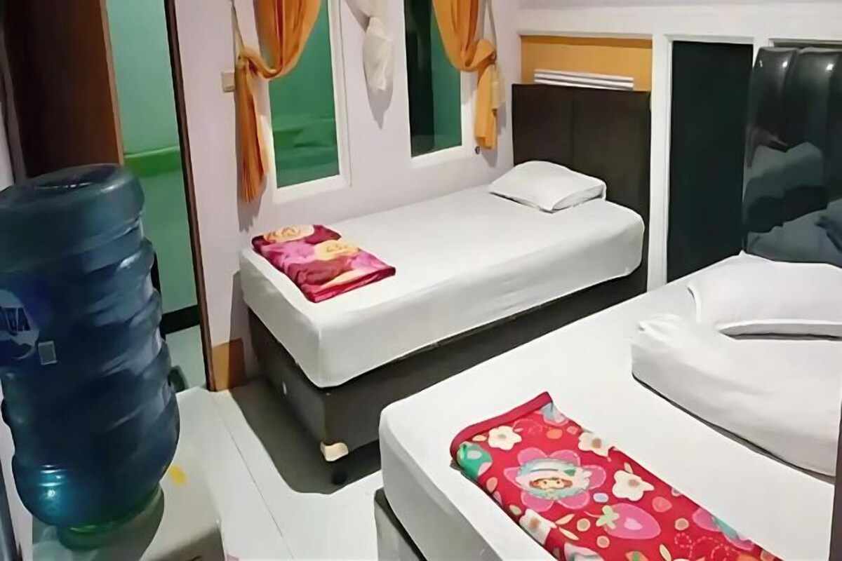 Quarto de hotel com duas camas de solteiro, um grande dispensador de água e uma pequena janela com cortinas.