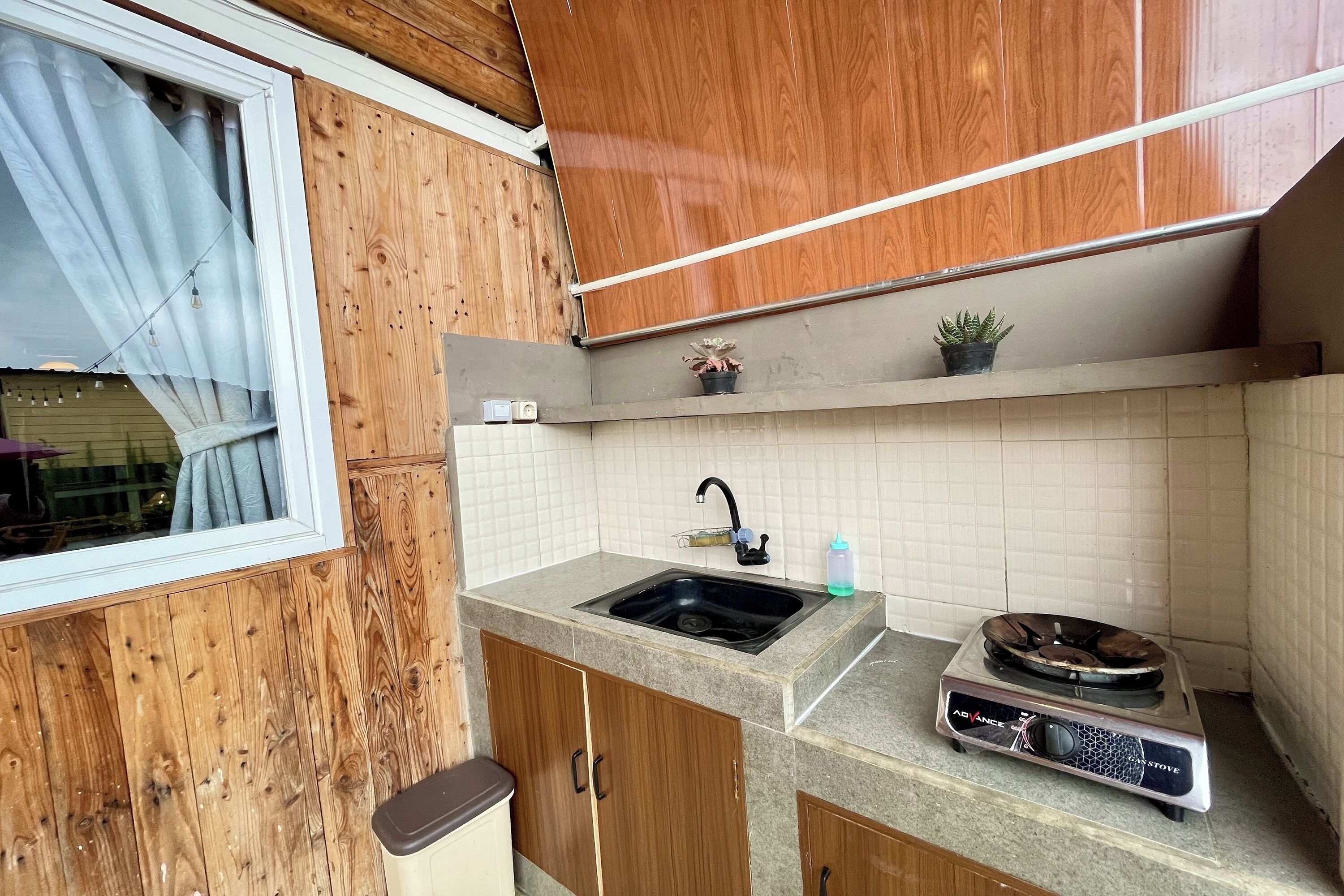 A compact kitchen with wooden cabinets, a sink, a gas stove, and a window with curtains.