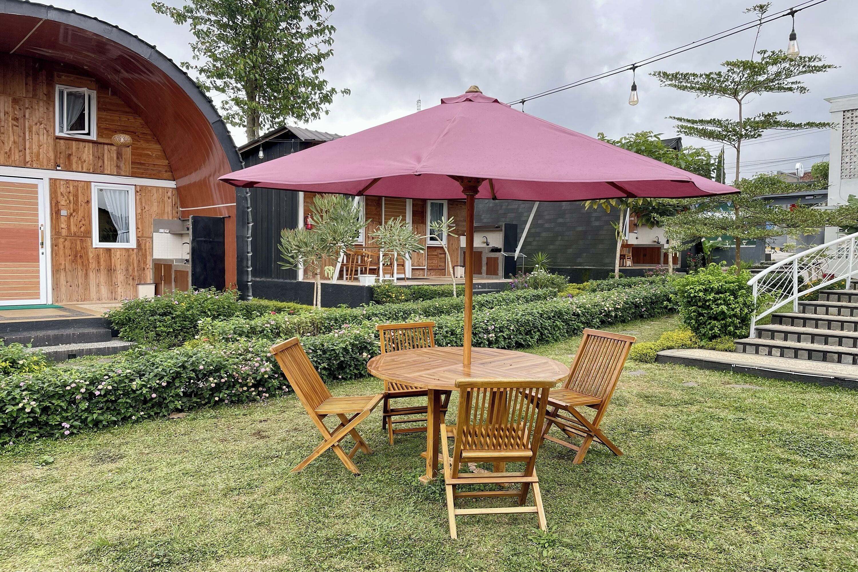 A wooden outdoor dining area with a table and chairs, surrounded by a well-maintained garden and a building with a curved facade.