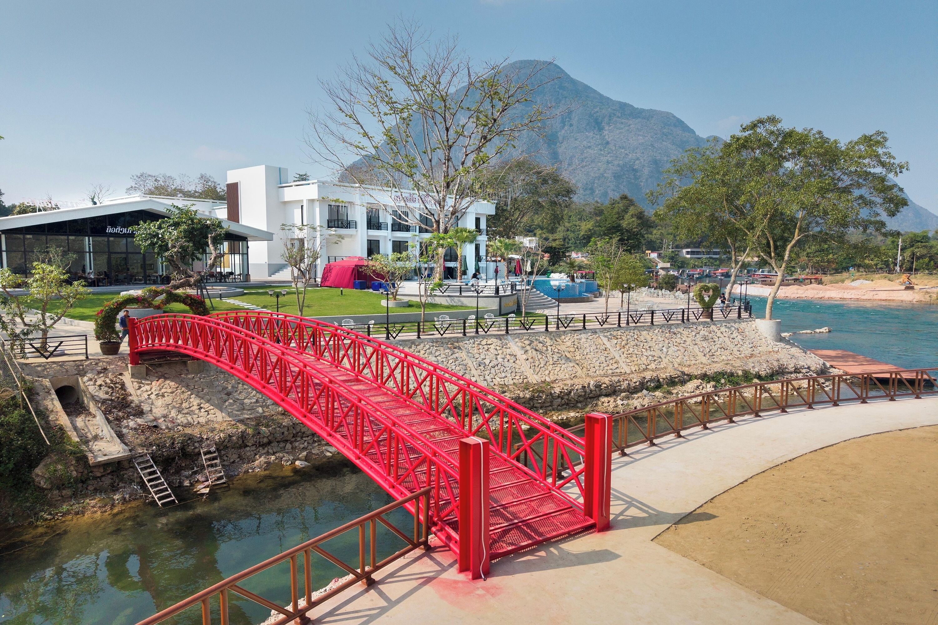 Un ponte rosso sopra un corso d'acqua, un edificio moderno con un cartello e montagne sullo sfondo.