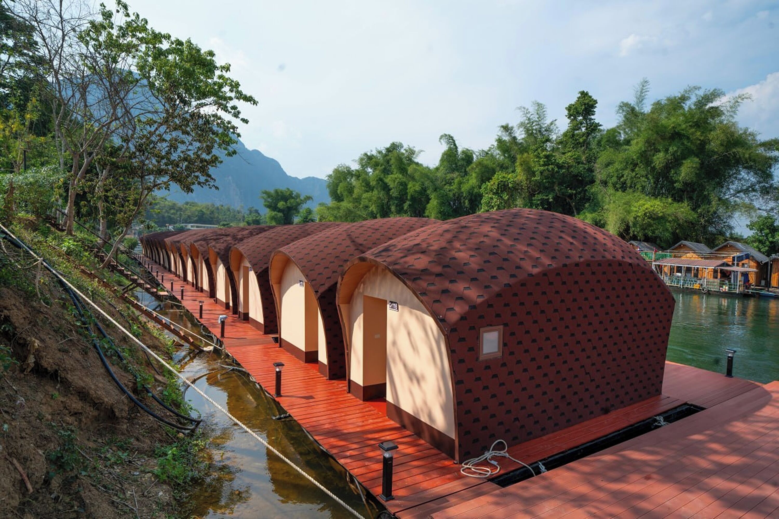 A row of dome-shaped cabins with a red roof and beige walls, lined up along a riverbank with a wooden walkway.