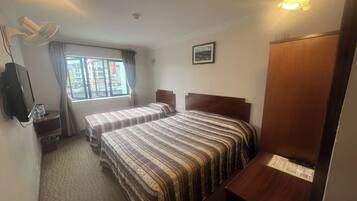 A hotel room with two beds, a desk, a chair, a TV, a fan, a window with curtains, and a picture on the wall.