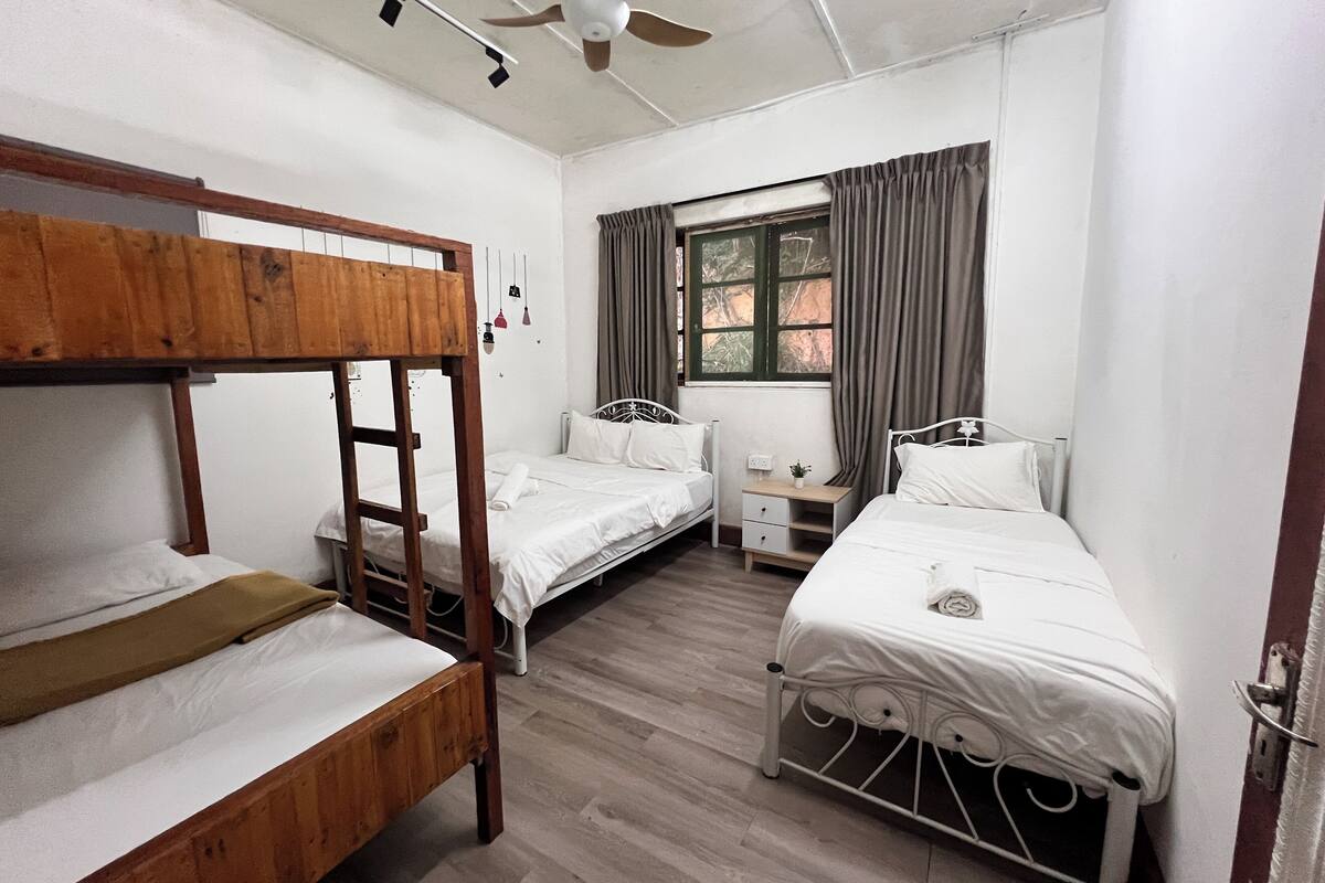 A dormitory room with bunk beds, a single bed, a window with curtains, a ceiling fan, and a small bedside table with a lamp.