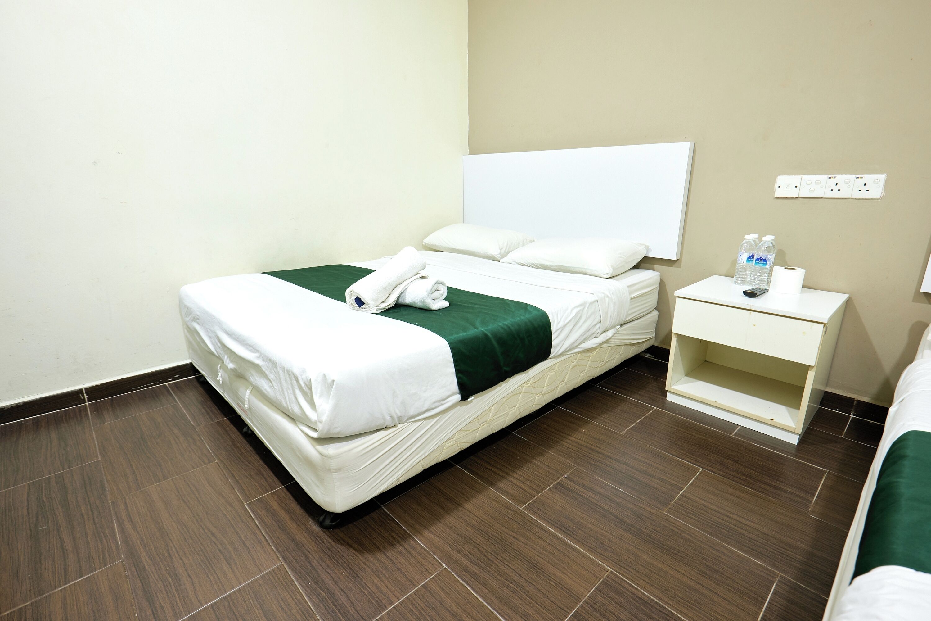 A neatly made bed with white linens and a green accent, a bedside table with a bottle and a tissue, and a power outlet on the wall.