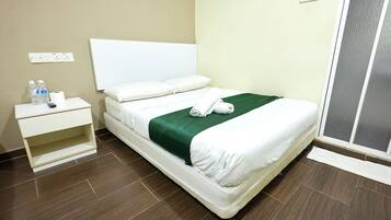 A neatly made bed with a green blanket and white linens, a bedside table with a remote control and bottled water, and a power outlet on the wall.
