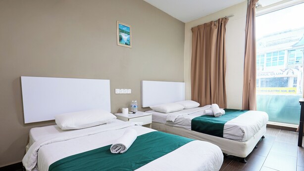 Habitación de hotel con dos camas, una ventana con cortinas, una mesita de noche con una botella y una caja de pañuelos, y un enchufe en la pared.