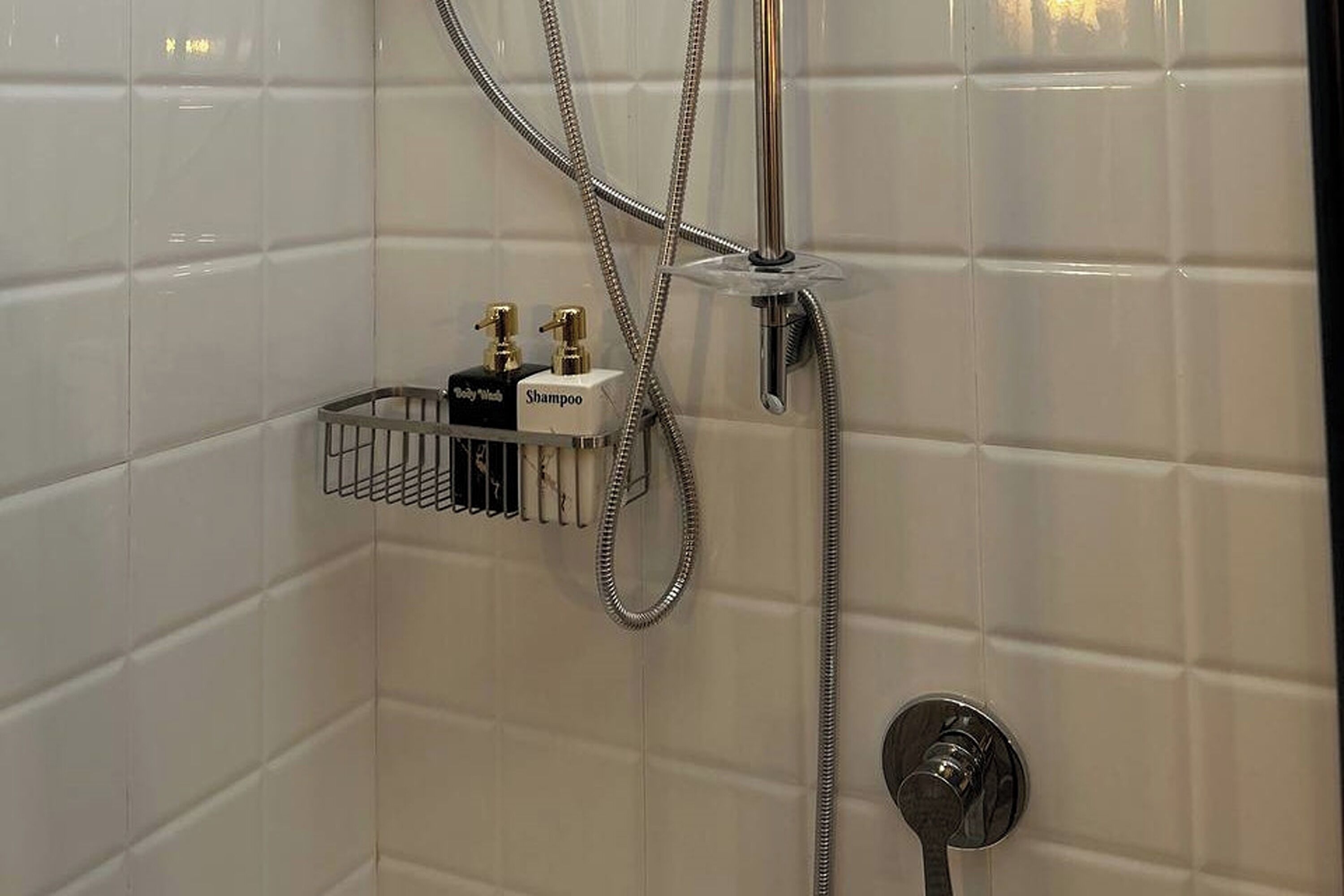 A bathroom with white tiled walls, a shower with a handheld showerhead, and a wall-mounted faucet. There is a small shelf with a shampoo bottle and a bottle of lotion.