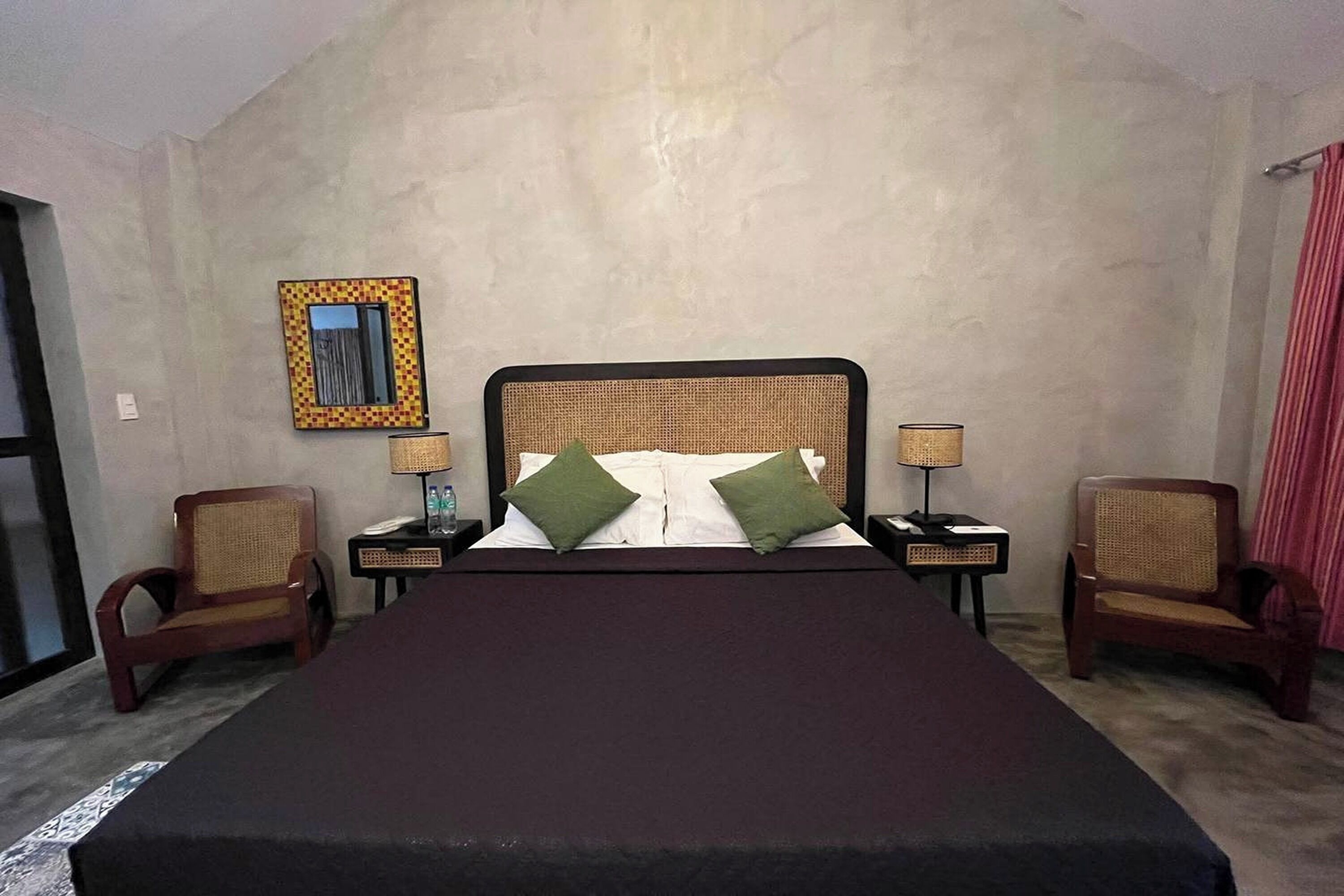 Habitación de hotel con una cama grande, dos mesitas de noche, dos sillones de mimbre, un espejo y una ventana.