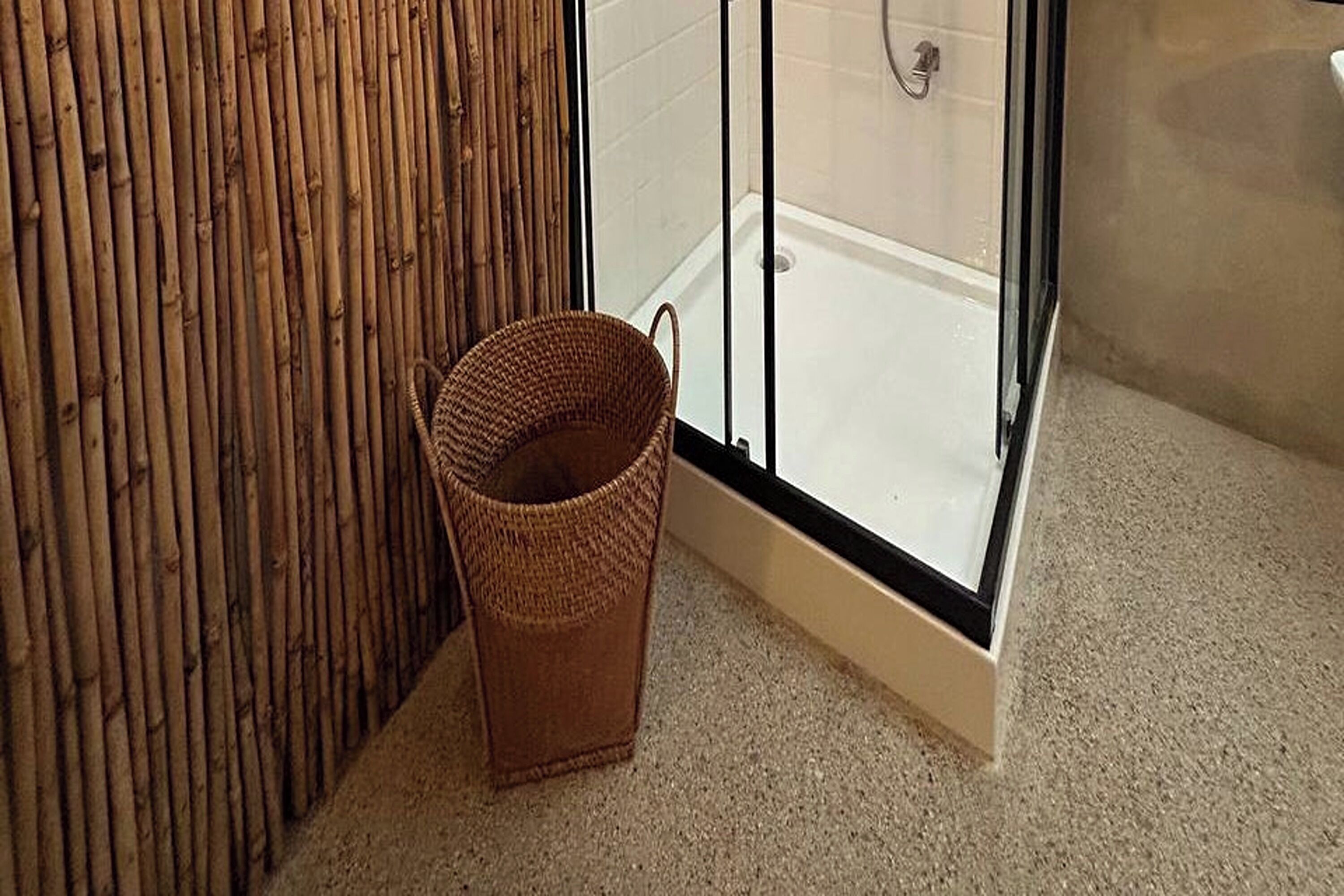 Un angolo di bagno con parete in bambù, box doccia in vetro e un cesto di vimini sul pavimento.