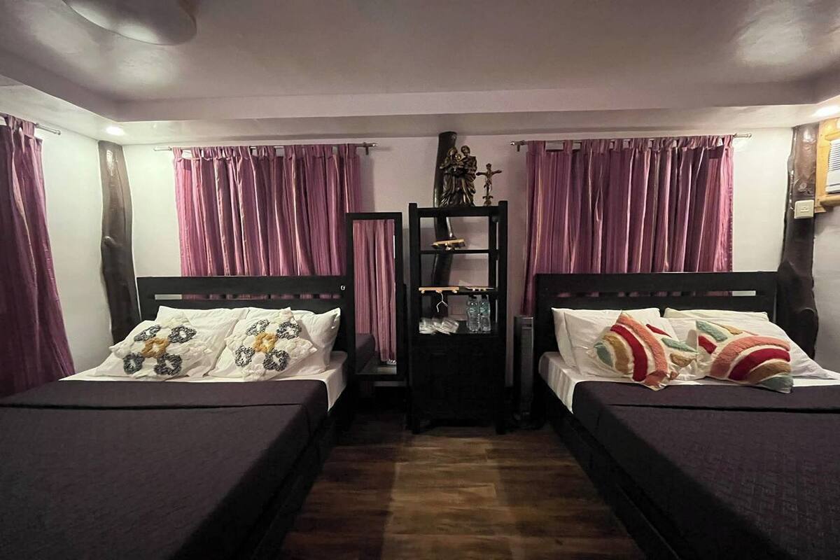 A room with two beds, each with a headboard and pillows. There is a shelf with various items on it, and the room has purple curtains.