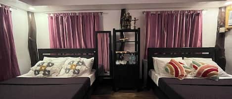 Una habitación con dos camas, cada una con cabecero y almohadas. Hay una estantería con varios objetos y cortinas moradas.
