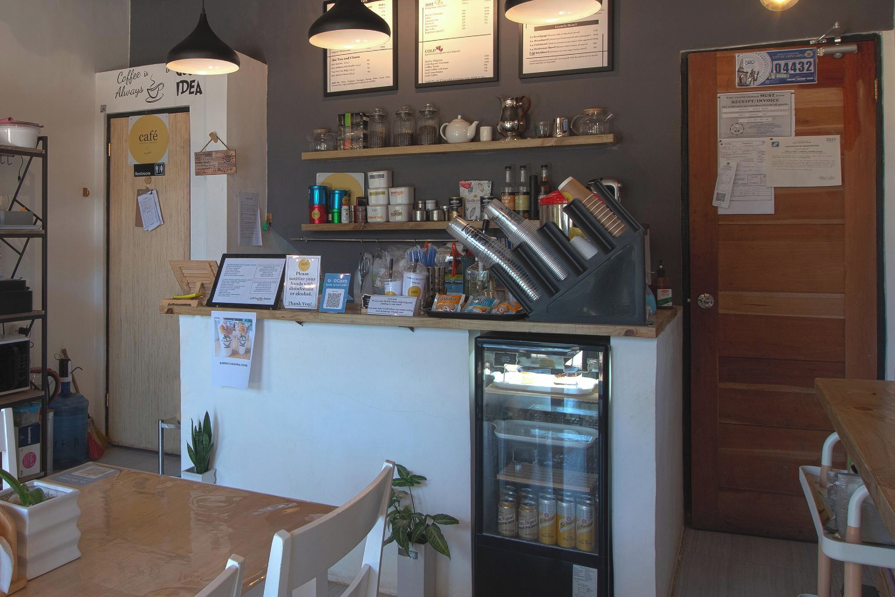 A café with a wooden counter, shelves with jars and bottles, a refrigerator, and a menu board.