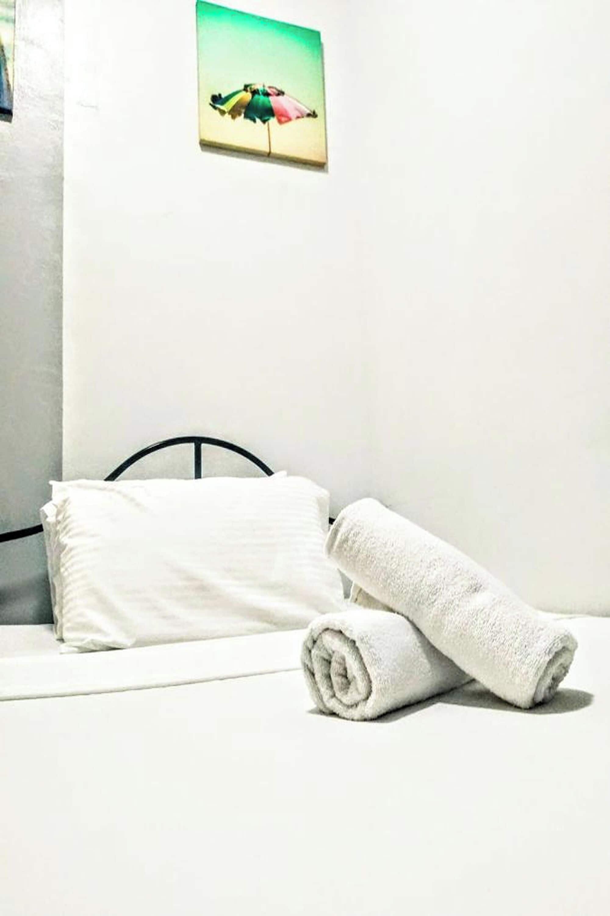 Un lit bien fait, avec du linge de lit blanc et une serviette pliée, une tête de lit en métal noir et un petit tableau représentant un parapluie coloré accroché au mur.