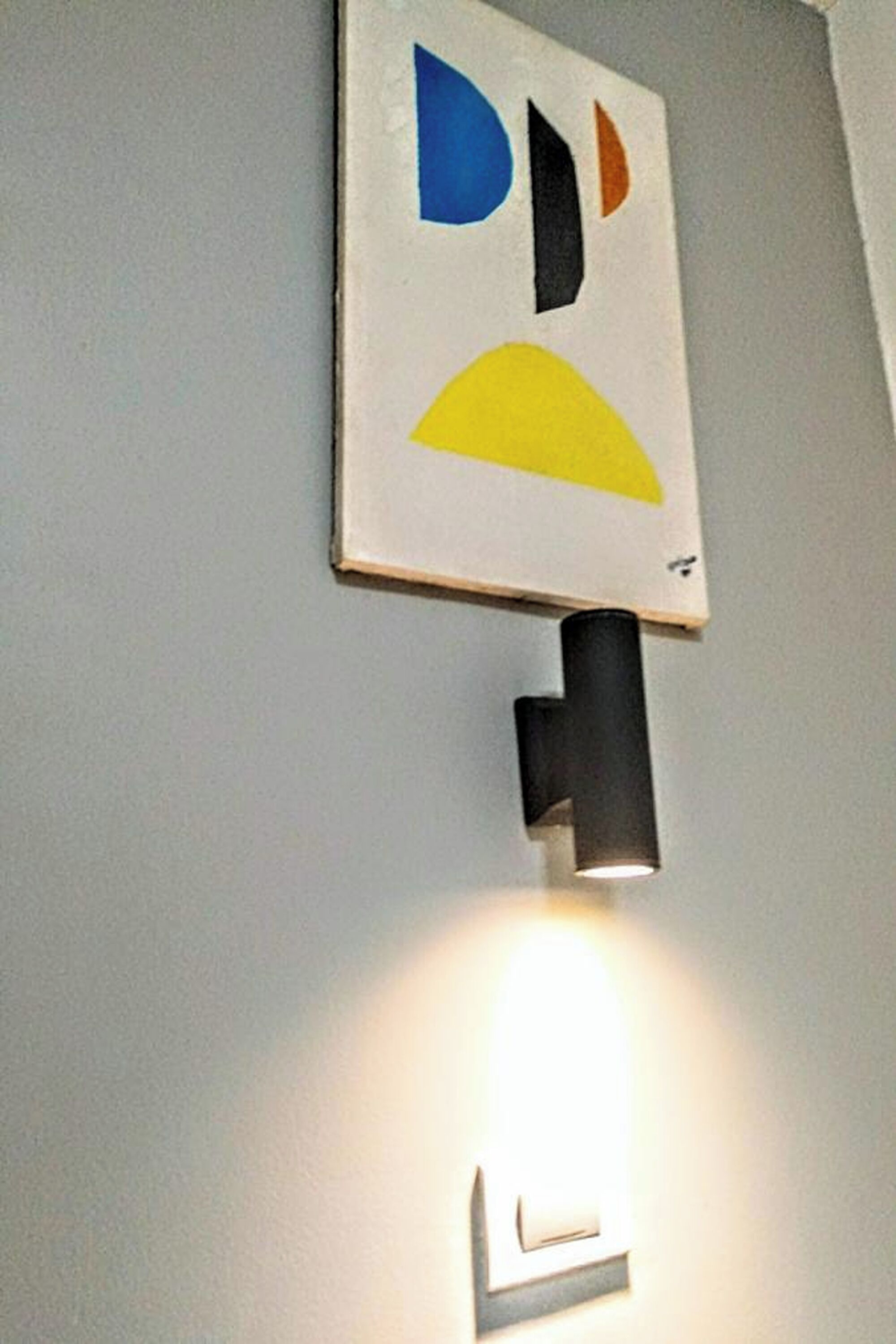 Seinään kiinnitetty valaisin, jonka yläpuolella on geometrinen taideteos.