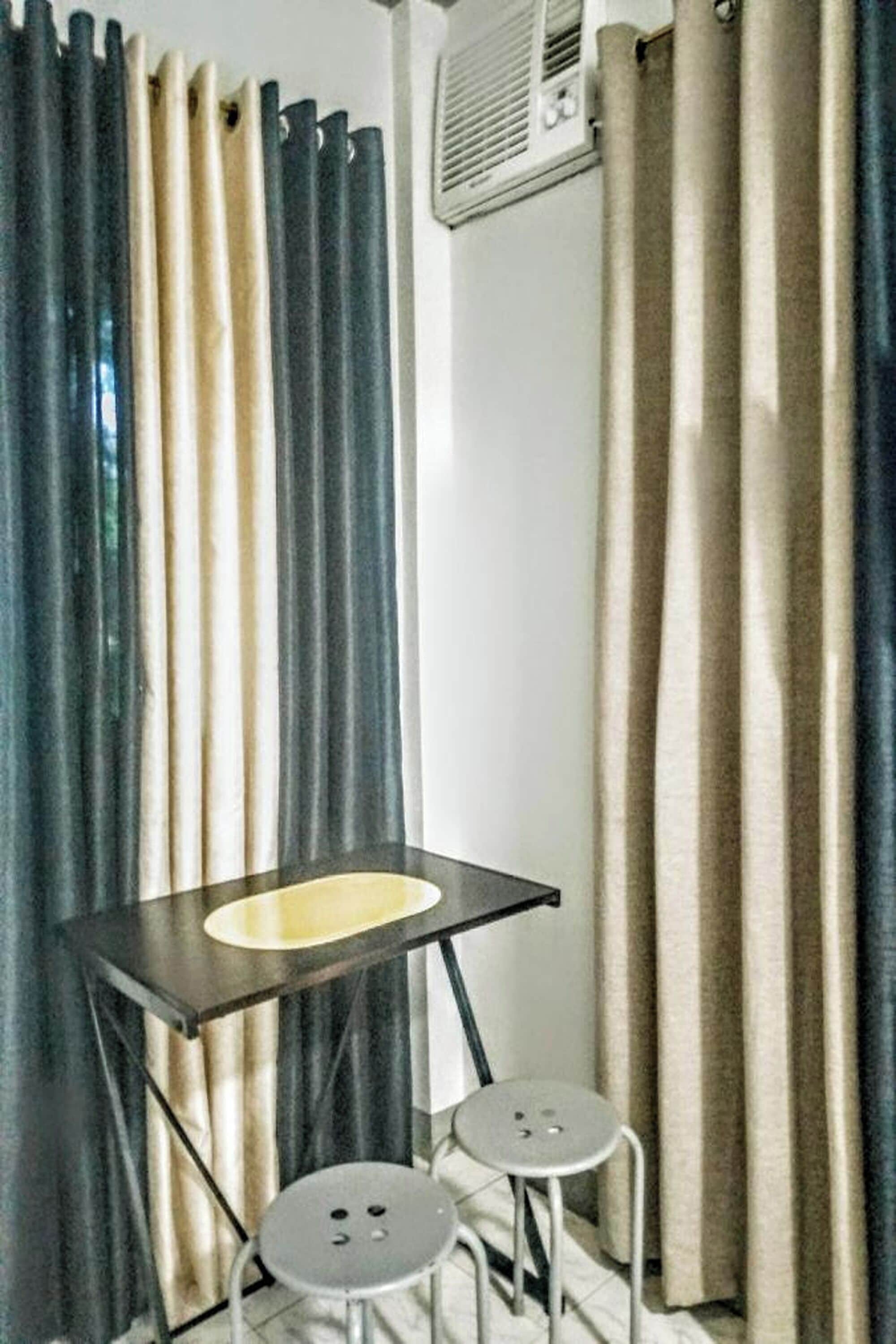 Una habitación pequeña con una mesa y dos taburetes, una ventana con cortinas y un equipo de aire acondicionado en la pared.