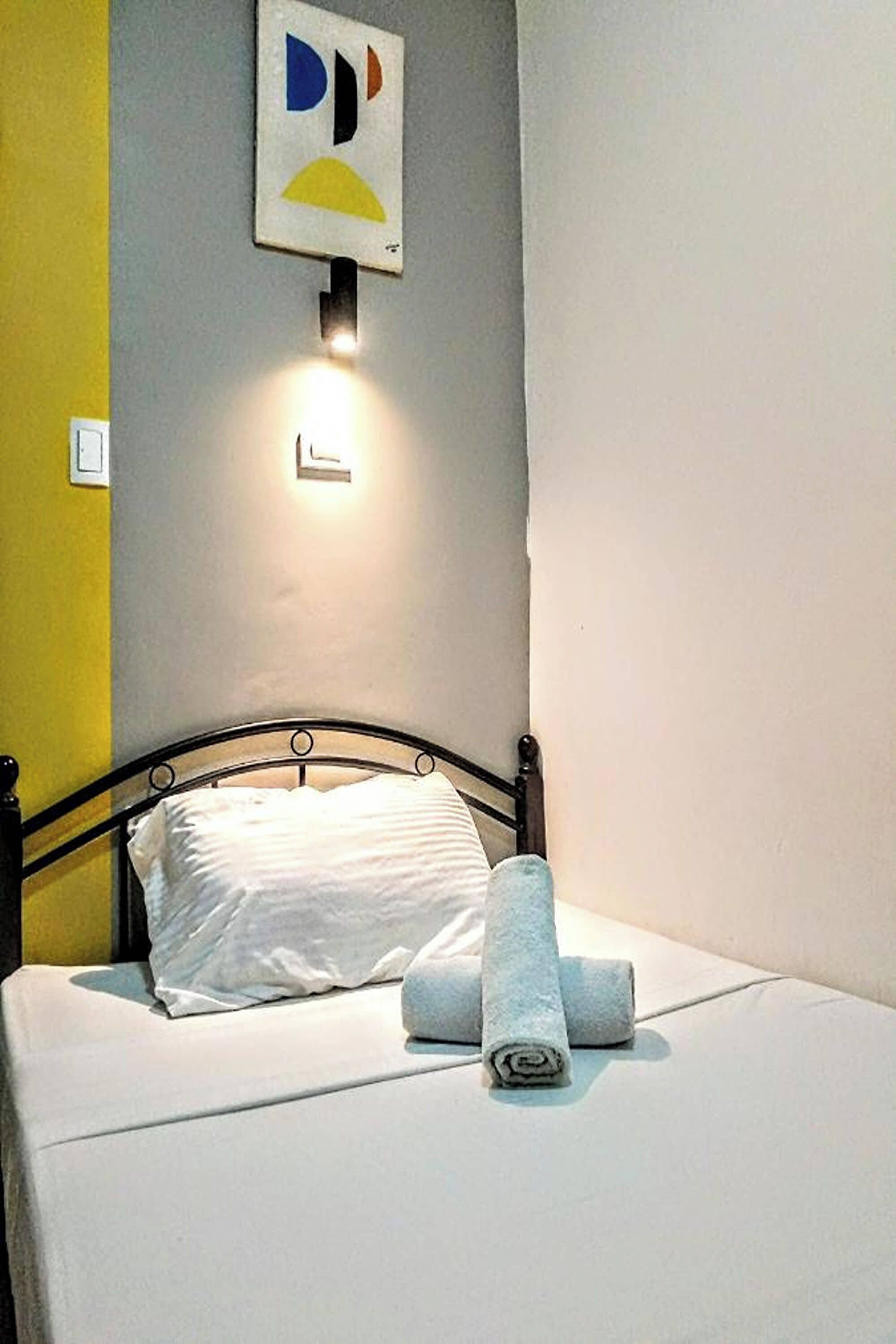 A neatly made bed with white linens and a rolled towel, a wall-mounted light fixture, and a partial view of a yellow wall.