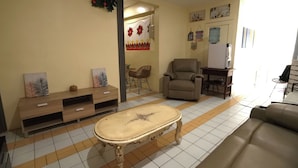 A living room with a sofa, a chair, a coffee table, and a TV stand. There is a small table with a chair and a water dispenser on the wall.