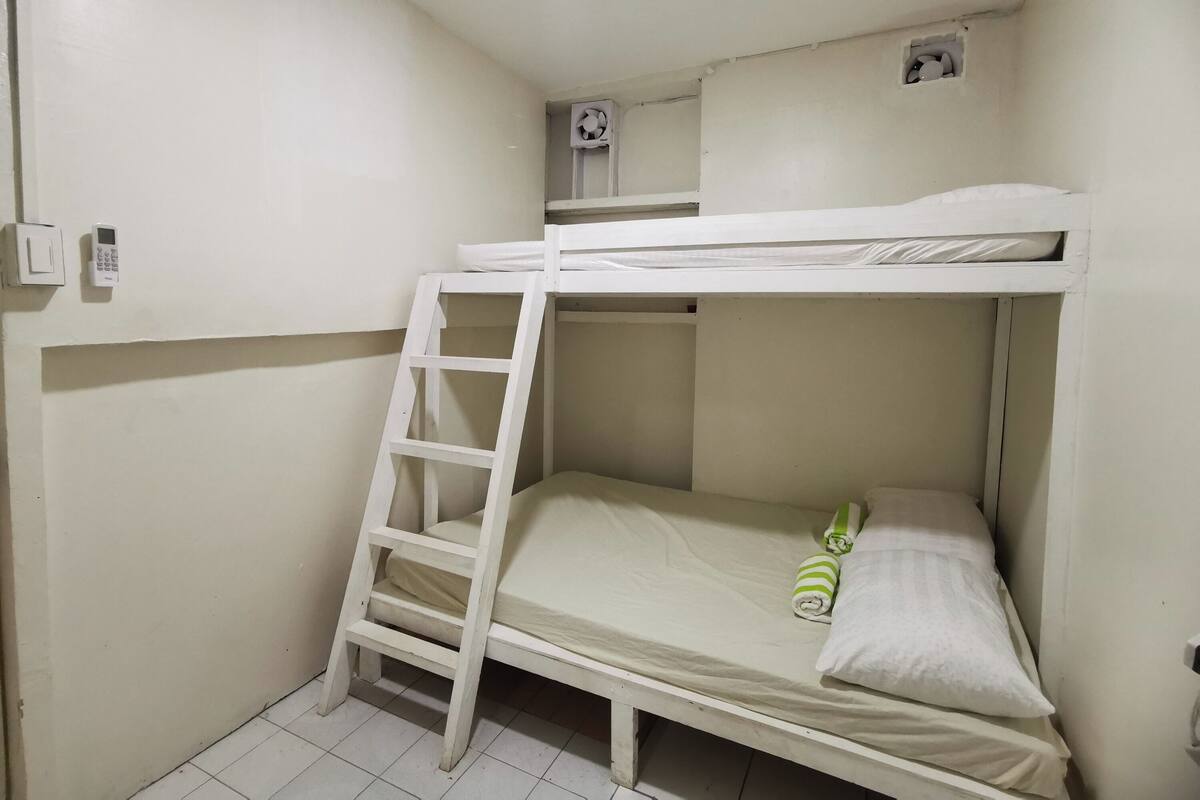 Habitación con literas, una escalera, un ventilador pequeño y un aplique en la pared.