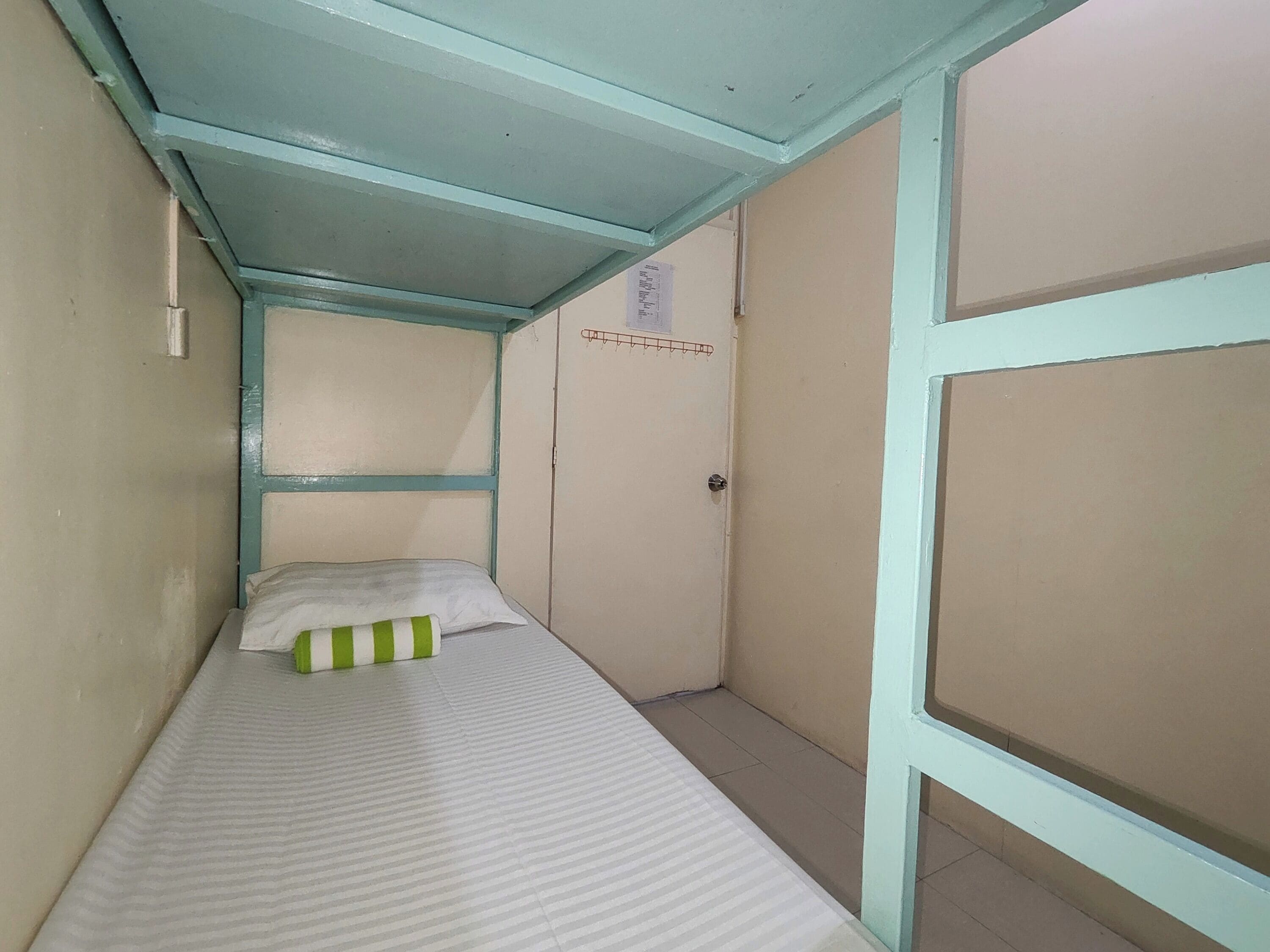 Ein schmales Einzelzimmer mit einem gestreiften Matratze und einem grün-weißen Kopfkissen. Das Zimmer hat eine hellblaue Decke und Wände, mit einer Tür auf der rechten Seite.