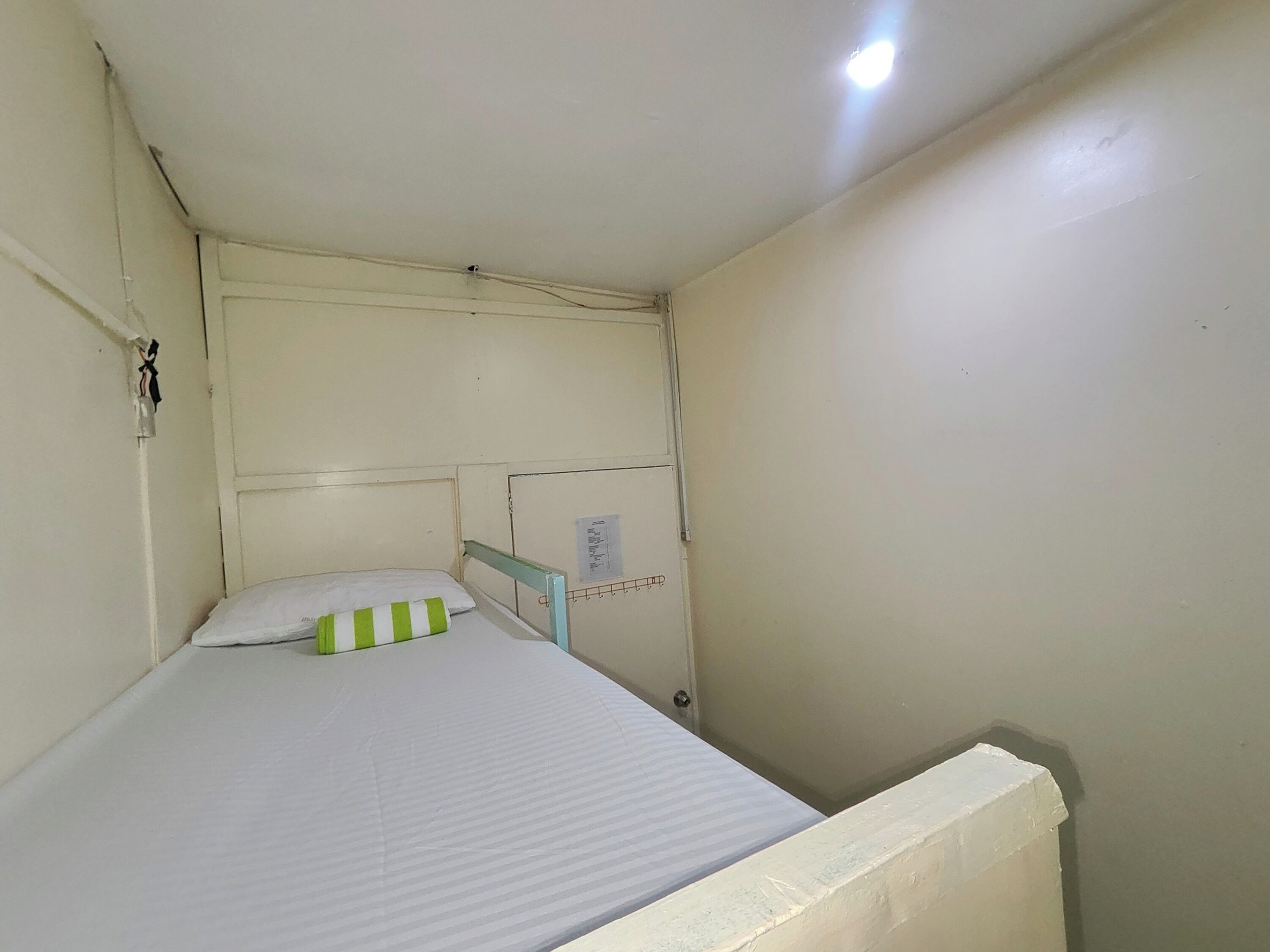 A small, single-bed room with a white mattress, a green and white striped pillow, and a white headboard.