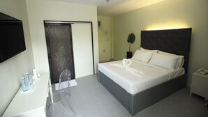 Quarto de hotel com cama, mesa de cabeceira com telefone, uma cadeira, escrivaninha com espelho e televisão na parede.