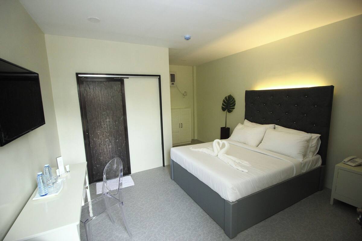 Ein Hotelzimmer mit einem Bett, einem Nachttisch mit Telefon, einem Sessel, einem Schreibtisch mit Spiegel und einem an der Wand montierten Fernseher.