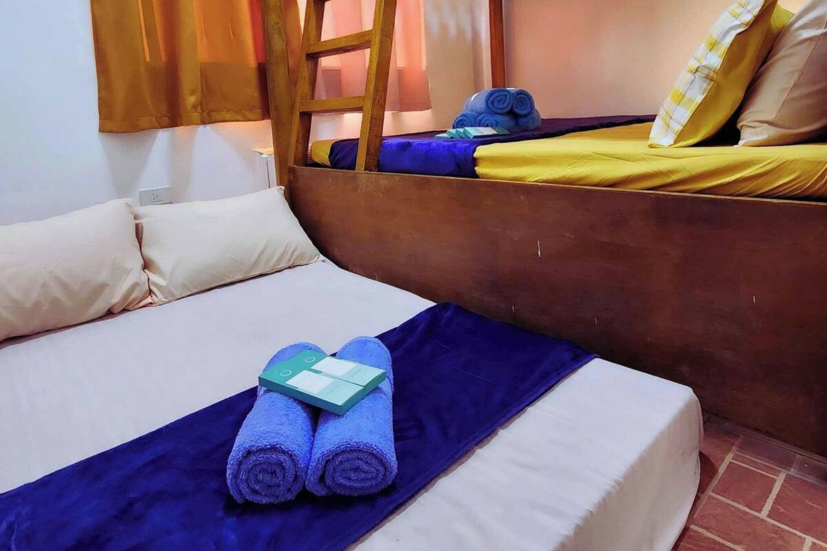 Une chambre avec un lit superposé, une échelle en bois, un lit recouvert d’une couverture bleue et agrémenté d’oreillers, et une fenêtre avec des rideaux.