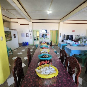 A long dining table with various dishes, chairs, and a kitchen area in the background. - Pertible Beach Resort By Zuzu (Anda)