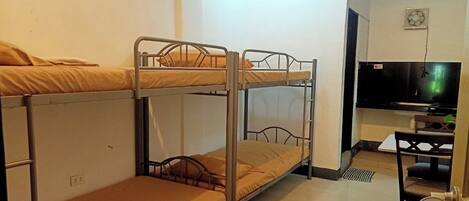 A dormitory room with bunk beds, a desk, a chair, a television, and a 'Now Available' sign.