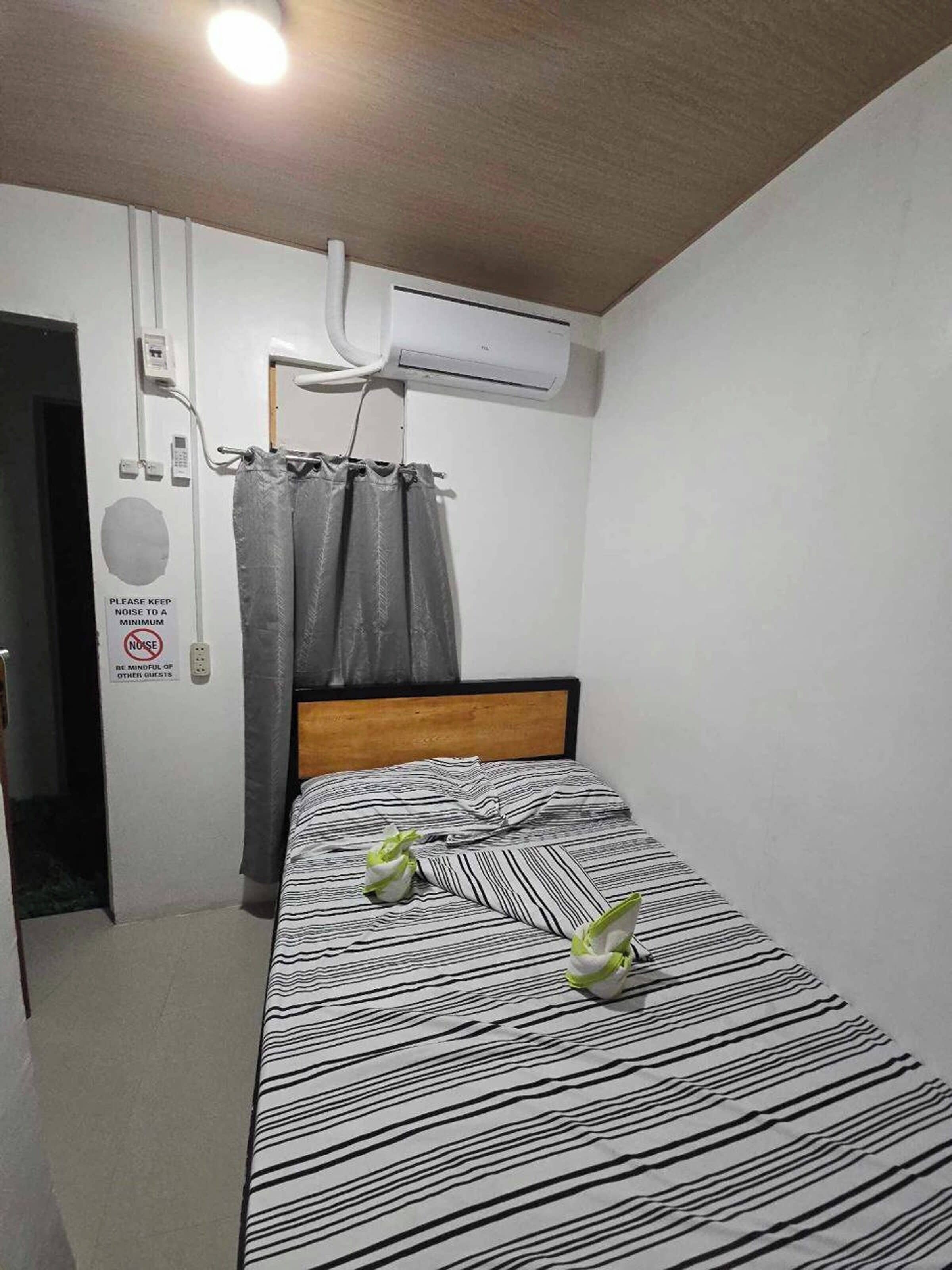 A small, neatly arranged room with a bed, a wall-mounted air conditioner, a door, and a sign requesting guests to keep noise to a minimum.