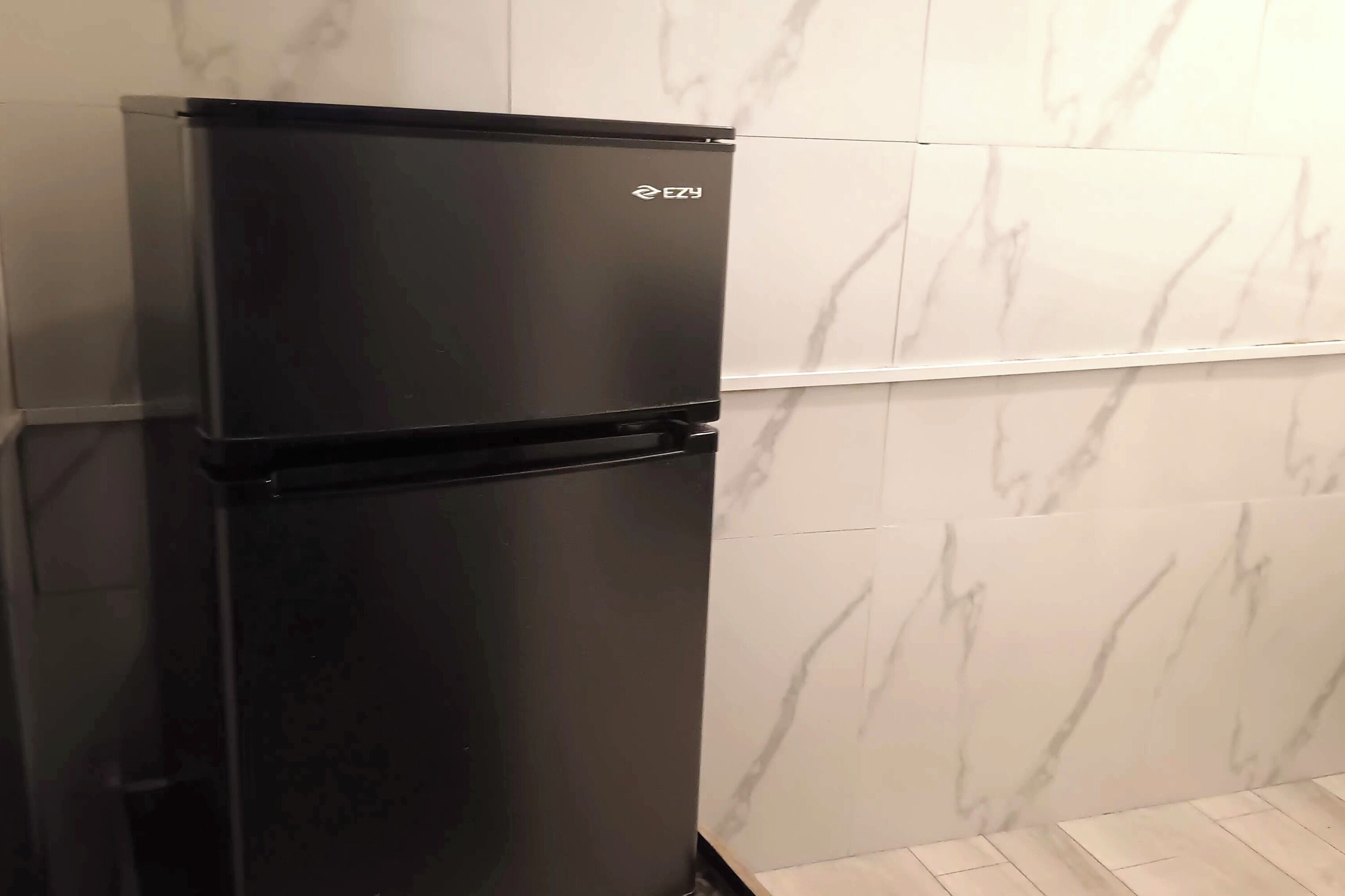 Un refrigerador negro con la marca 'EZY' en una cocina con baldosas de mármol blanco y piso de madera.