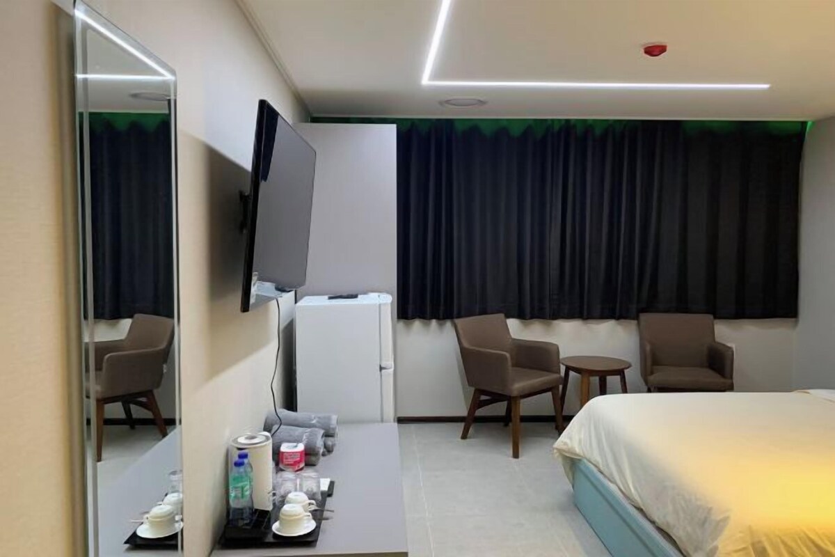A hotel room with a bed, a desk with a flat-screen TV, two chairs, a small table, and a window with curtains.