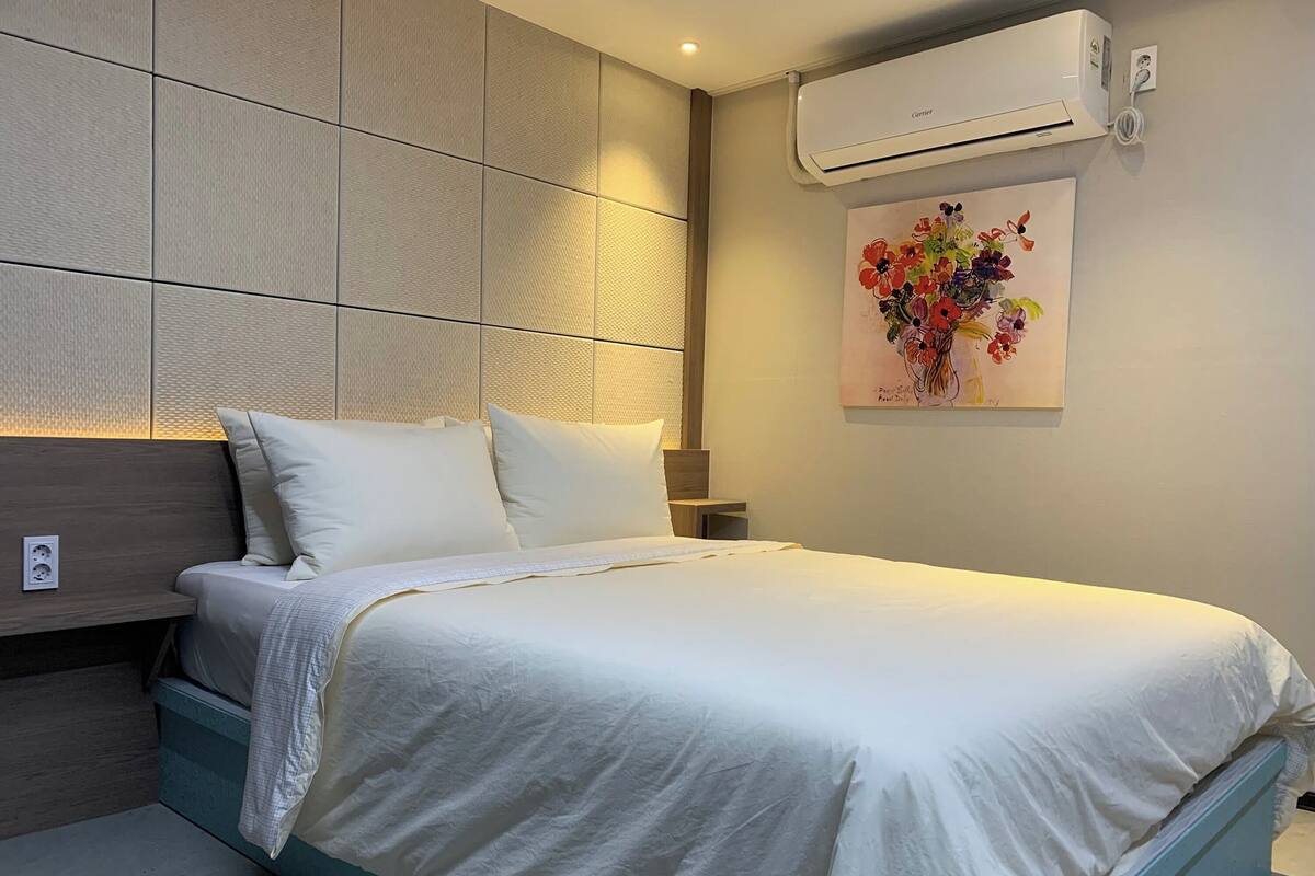A modern bedroom with a large bed, a headboard with a textured panel, an air conditioning unit, and a floral artwork on the wall.
