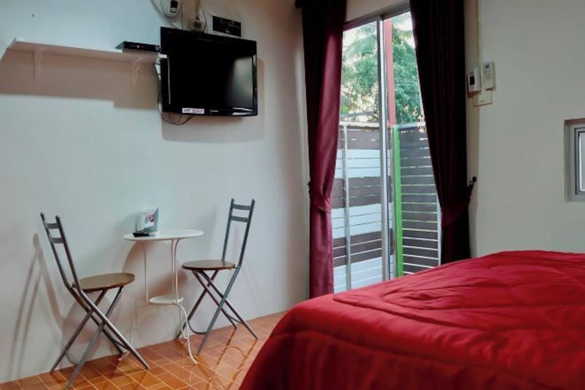 Una habitación con cama, televisor montado en la pared, una mesita con dos sillas y una ventana con cortinas.