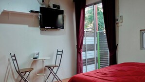 A room with a bed, a TV mounted on the wall, a small table with chairs, and a window with curtains.