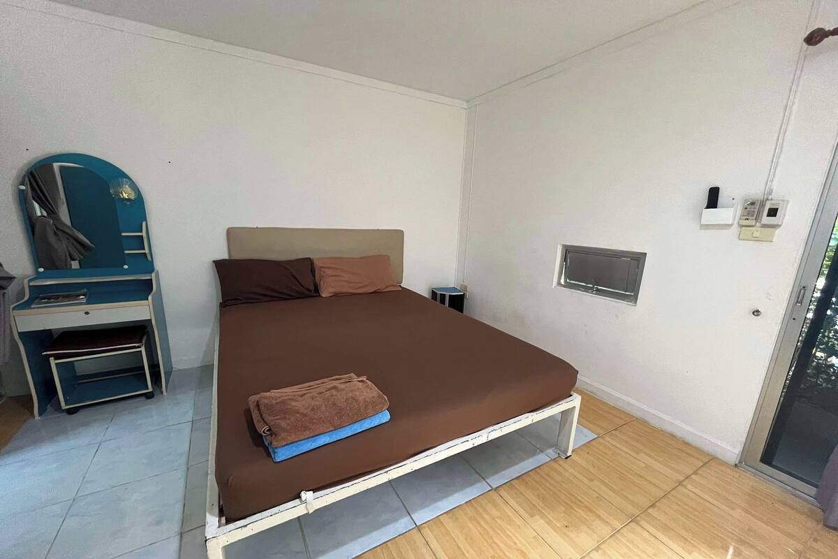 Uma cama de solteiro com colchão castanho e manta azul, uma pequena mesa de cabeceira, uma escrivaninha com cadeira e uma janela com vista para árvores.