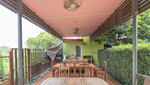 An outdoor patio with a wooden table and chairs, a ceiling fan, and a view of a green building in the background.