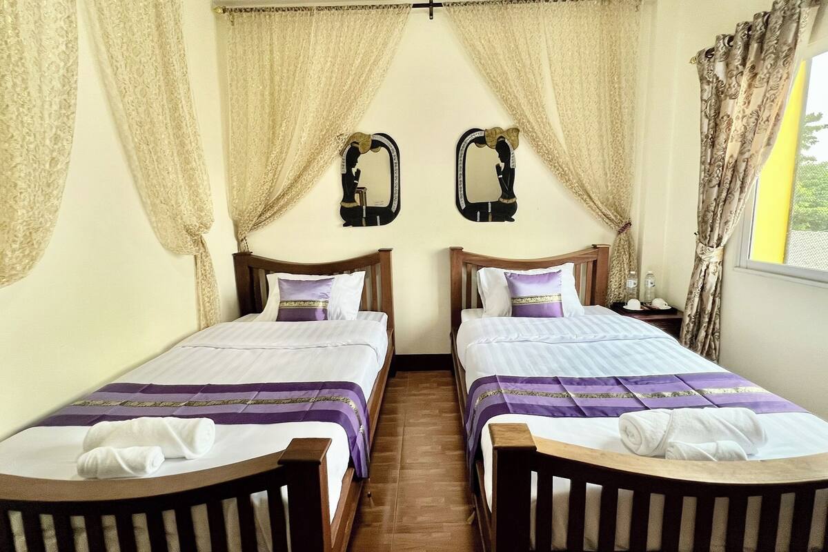 A room with two wooden beds, each with a headboard, white linens, and purple accents. There are two wall-mounted mirrors and a window with curtains.