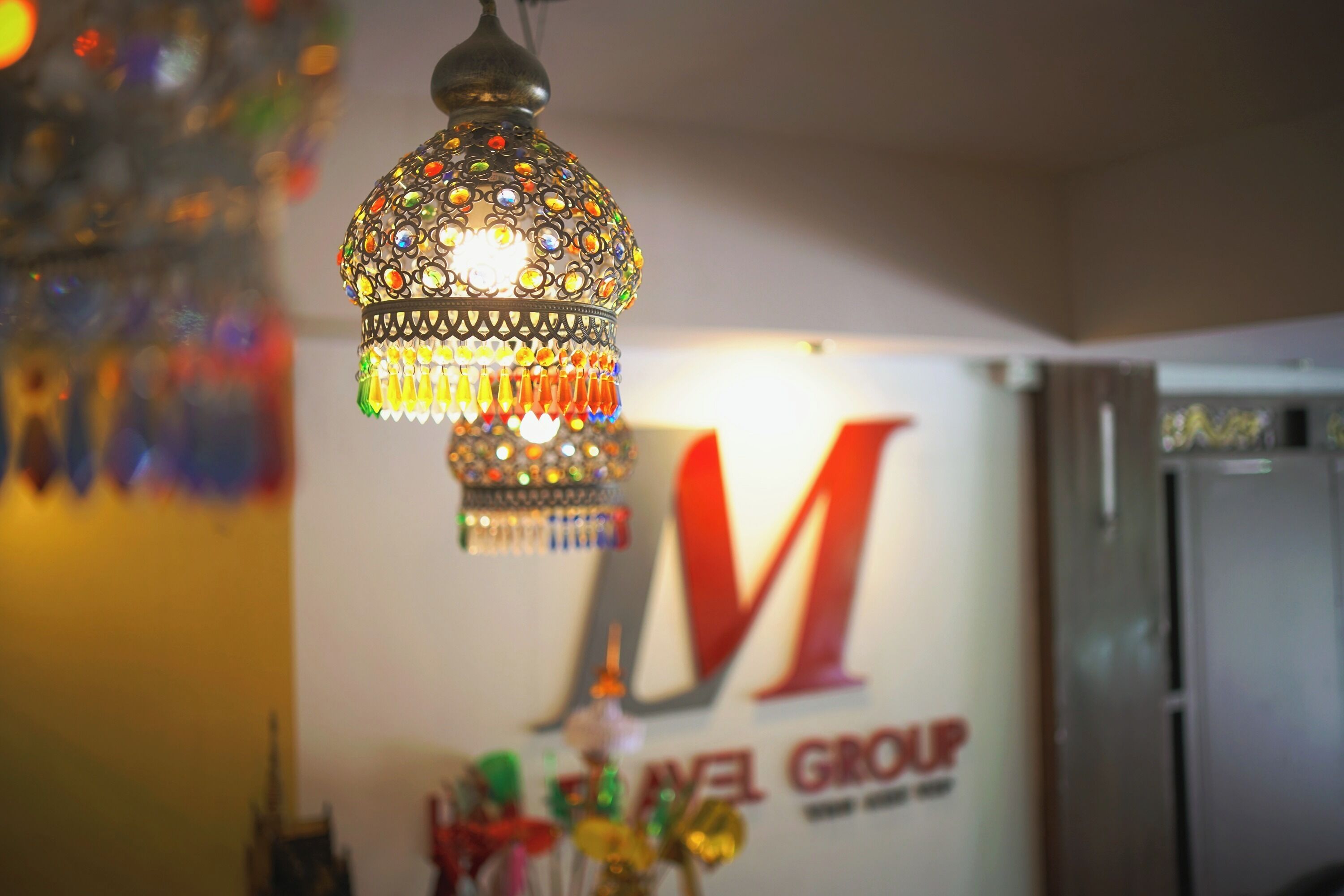 A hanging decorative lamp with intricate designs and colorful lights, a sign with the letters "M" and "G" for "Motel Group," and a glimpse of a room with a door and a refrigerator.