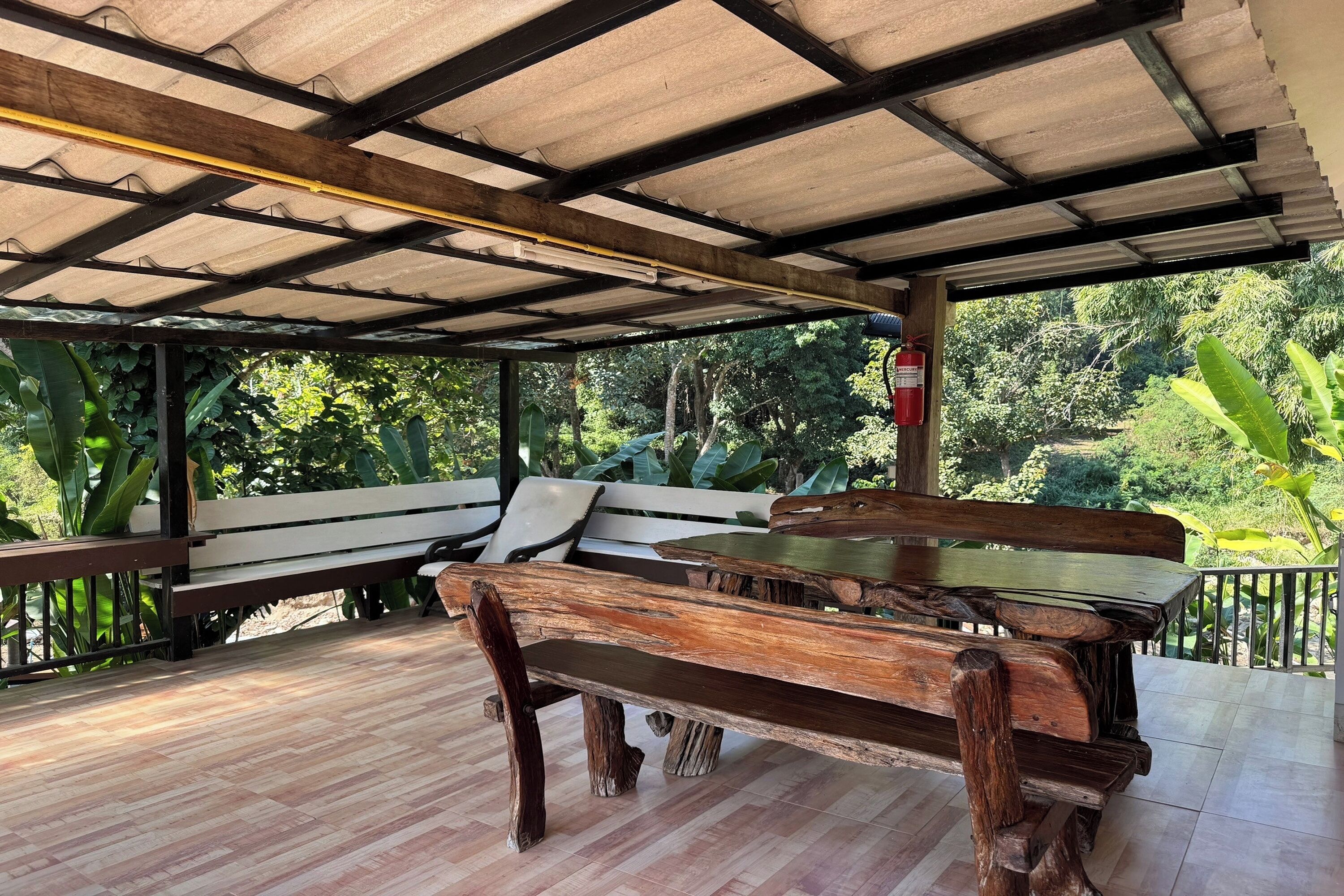 An outdoor covered patio with wooden benches and tables, surrounded by lush greenery.