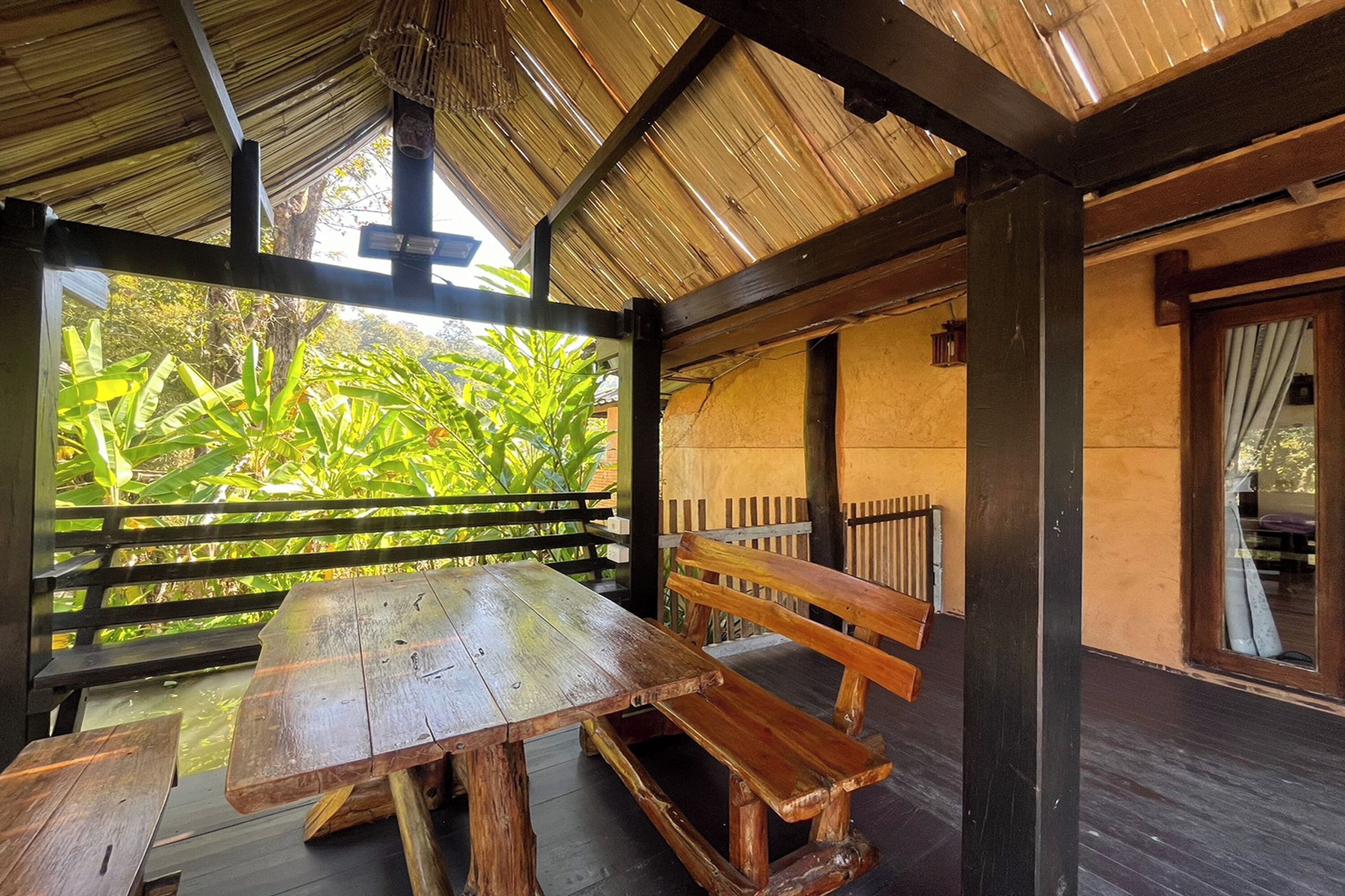 A wooden bench and table under a thatched roof, with a view of lush greenery outside.