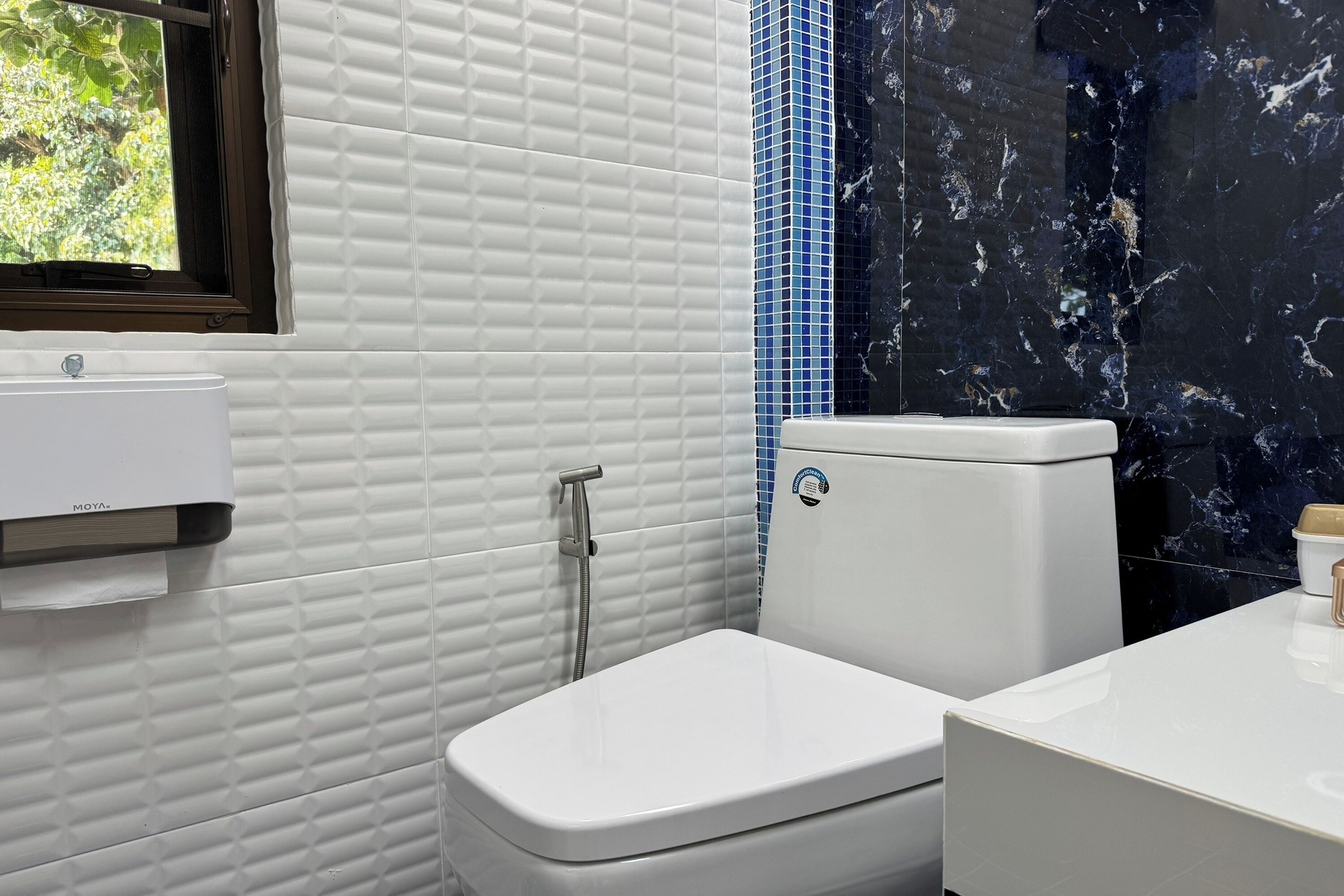 Une salle de bain avec des toilettes blanches, un lavabo blanc et une fenêtre donnant sur un paysage verdoyant.