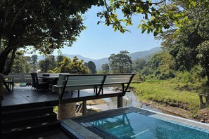 Un área de piscina con cubierta de madera y mobiliario exterior, rodeada de exuberante vegetación y vistas a colinas distantes.