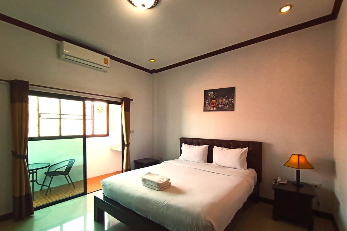 A hotel room with a bed, a bedside table, a lamp, a window with curtains, and a wall-mounted air conditioner.