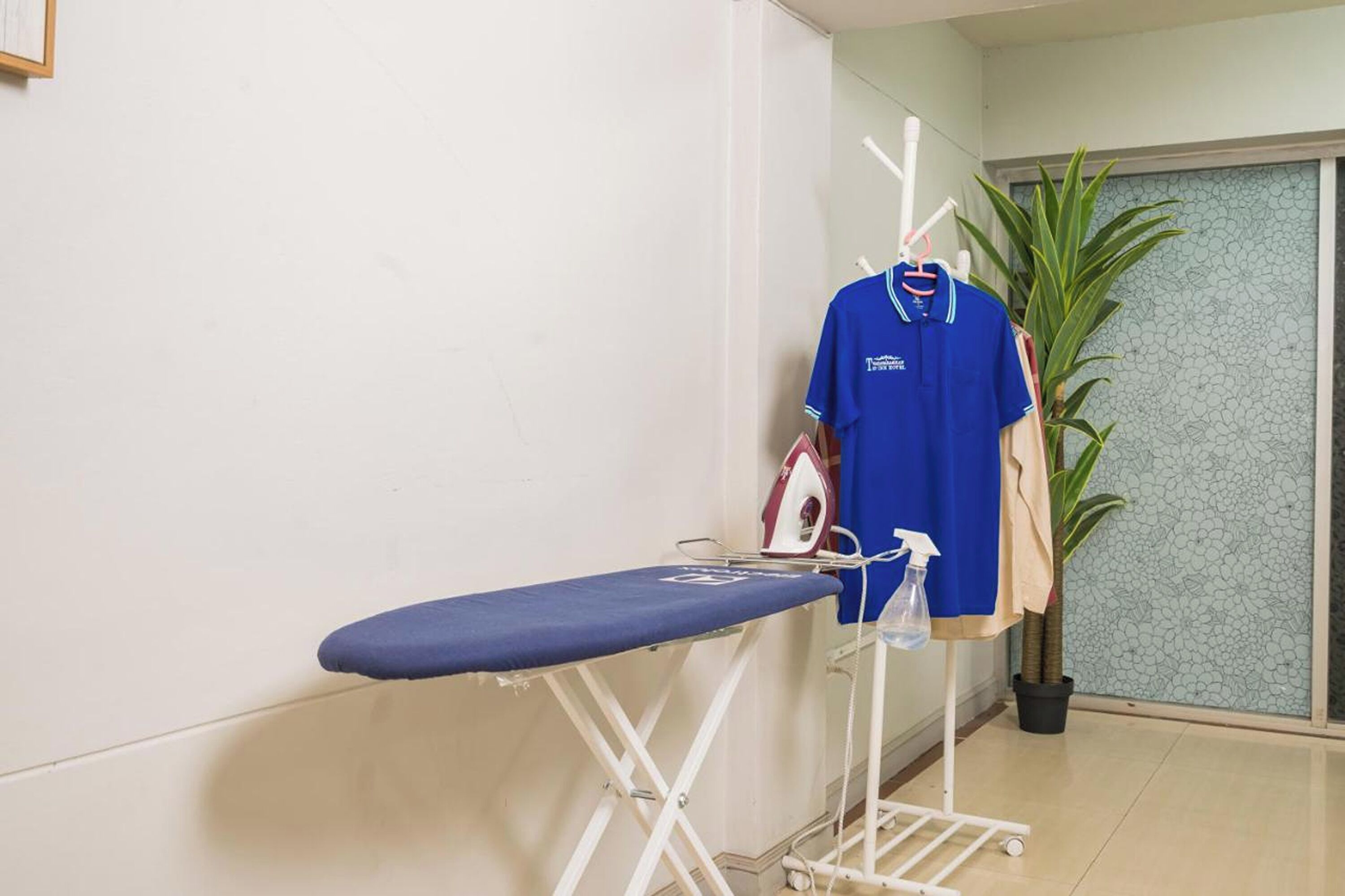 A room with a blue ironing board, an iron, and a hanging blue shirt.