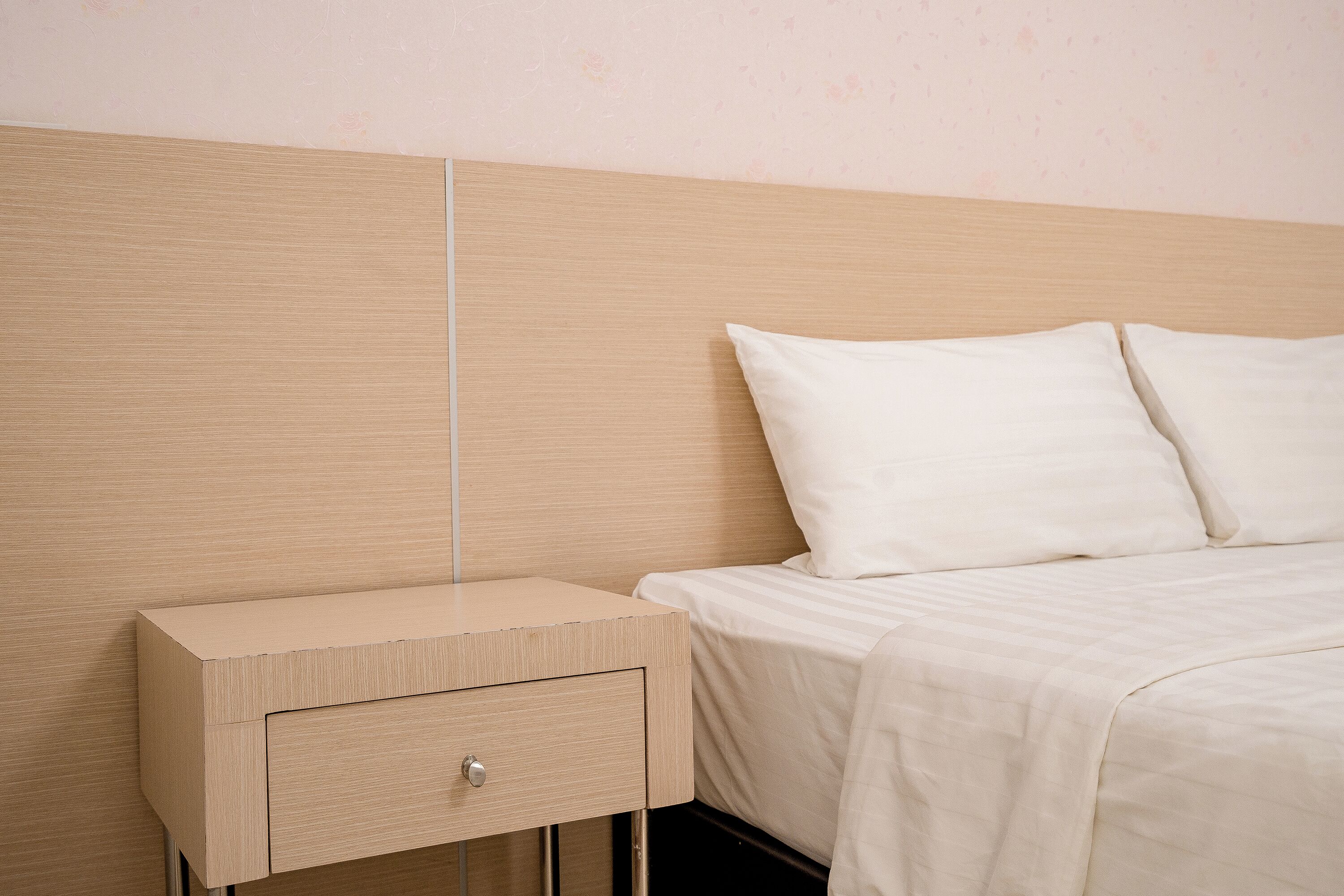 Un lit bien fait, avec du linge de lit blanc, une table de chevet en bois dotée d’un tiroir et un mur sobre.